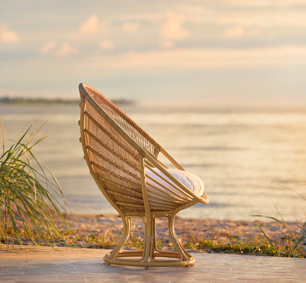 This is a lifestyle image of Luna Exterior Lounge Chair in natural aluminium artfibre. Placed on the shore at golden sunset with ocean behind.