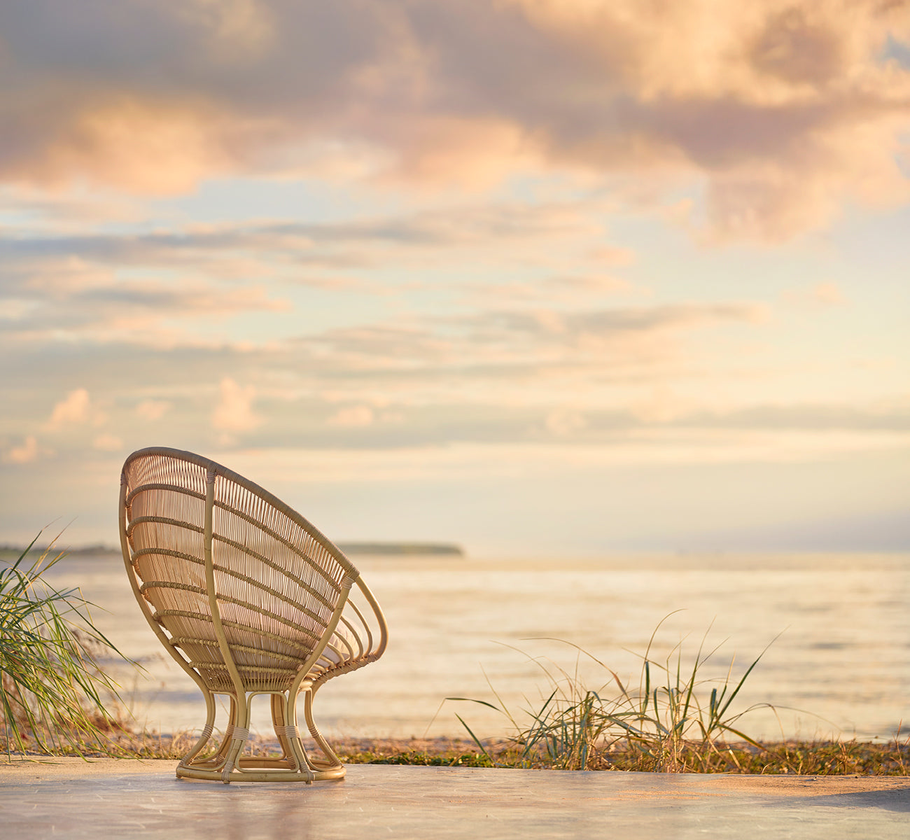 This is a lifestyle image of Luna Exterior Lounge Chair in natural aluminium artfibre. Seen from behind on a beach at sunset with a pastel sky.