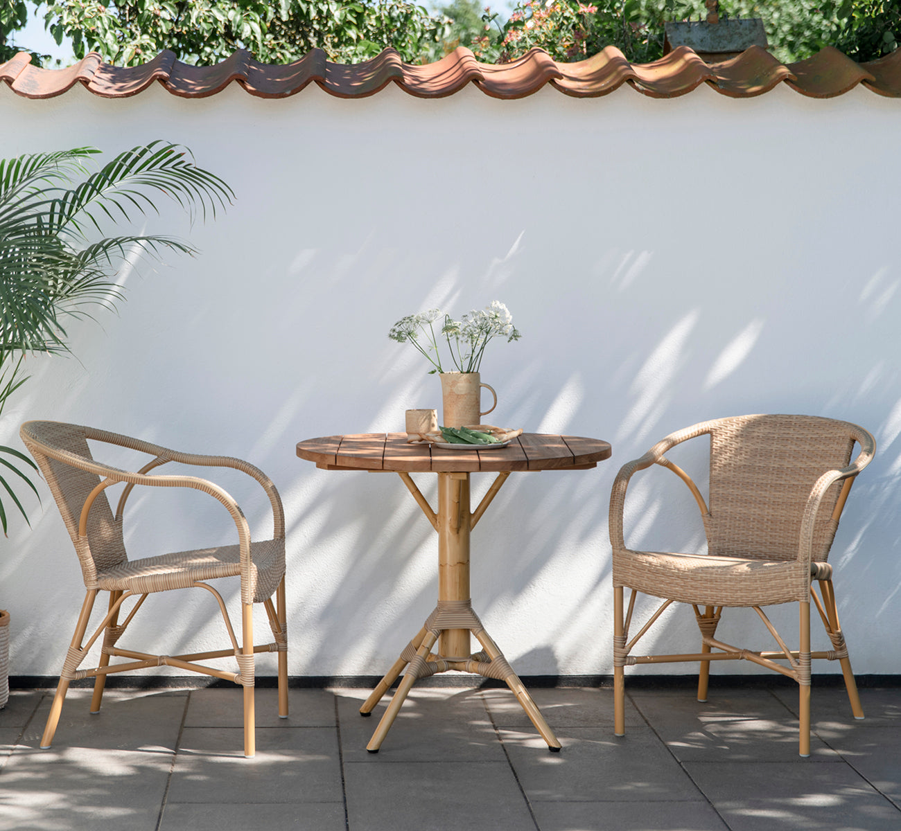This is a lifestyle image of Madeleine Exterior Dining Chair in almond aluminium frame natural ArtFibre. Courtyard bistro with round teak table.