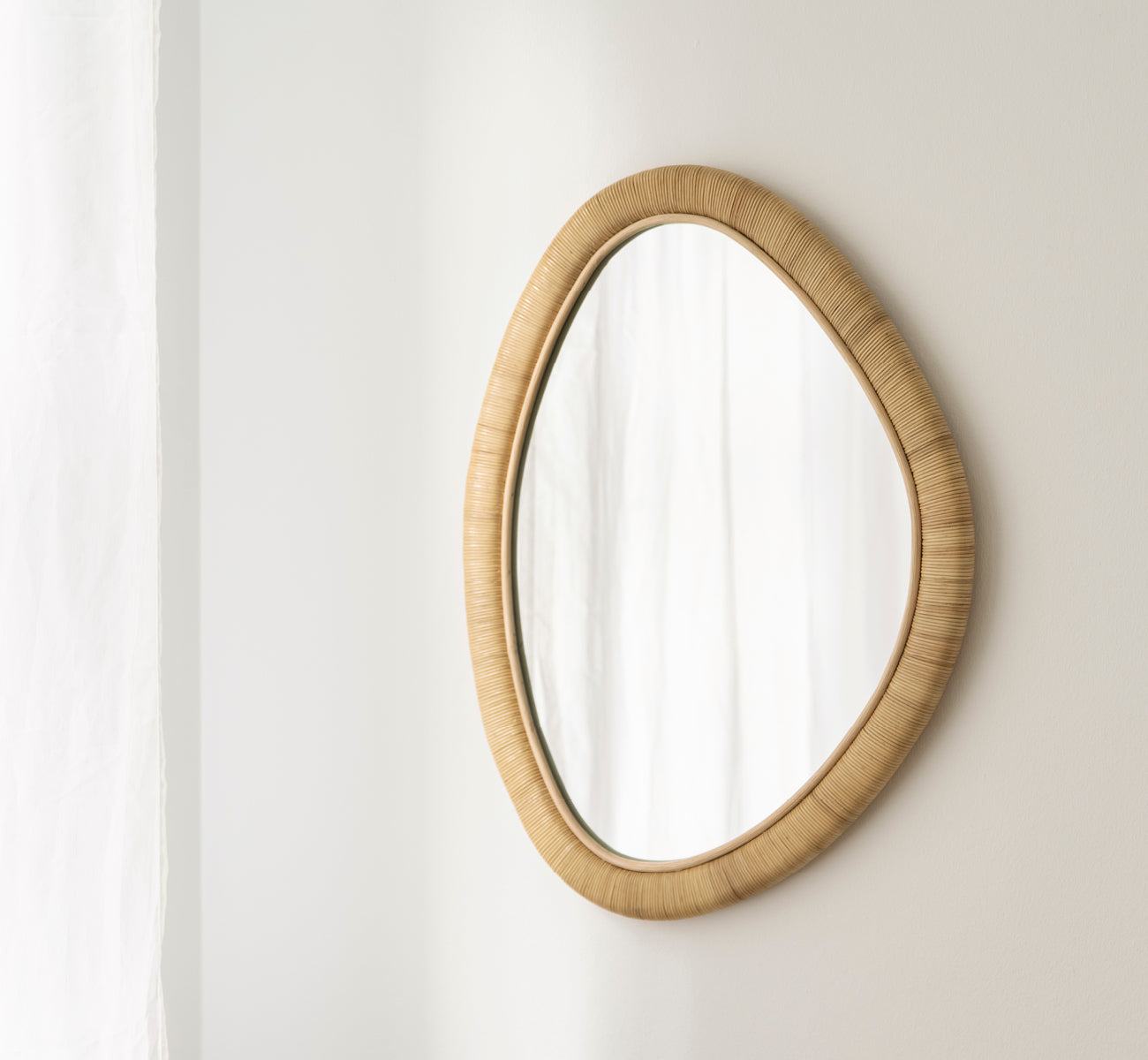 This is a lifestyle image of Malou Mirror in natural rattan. Organic oval shape mounted on a soft white wall with curtain.