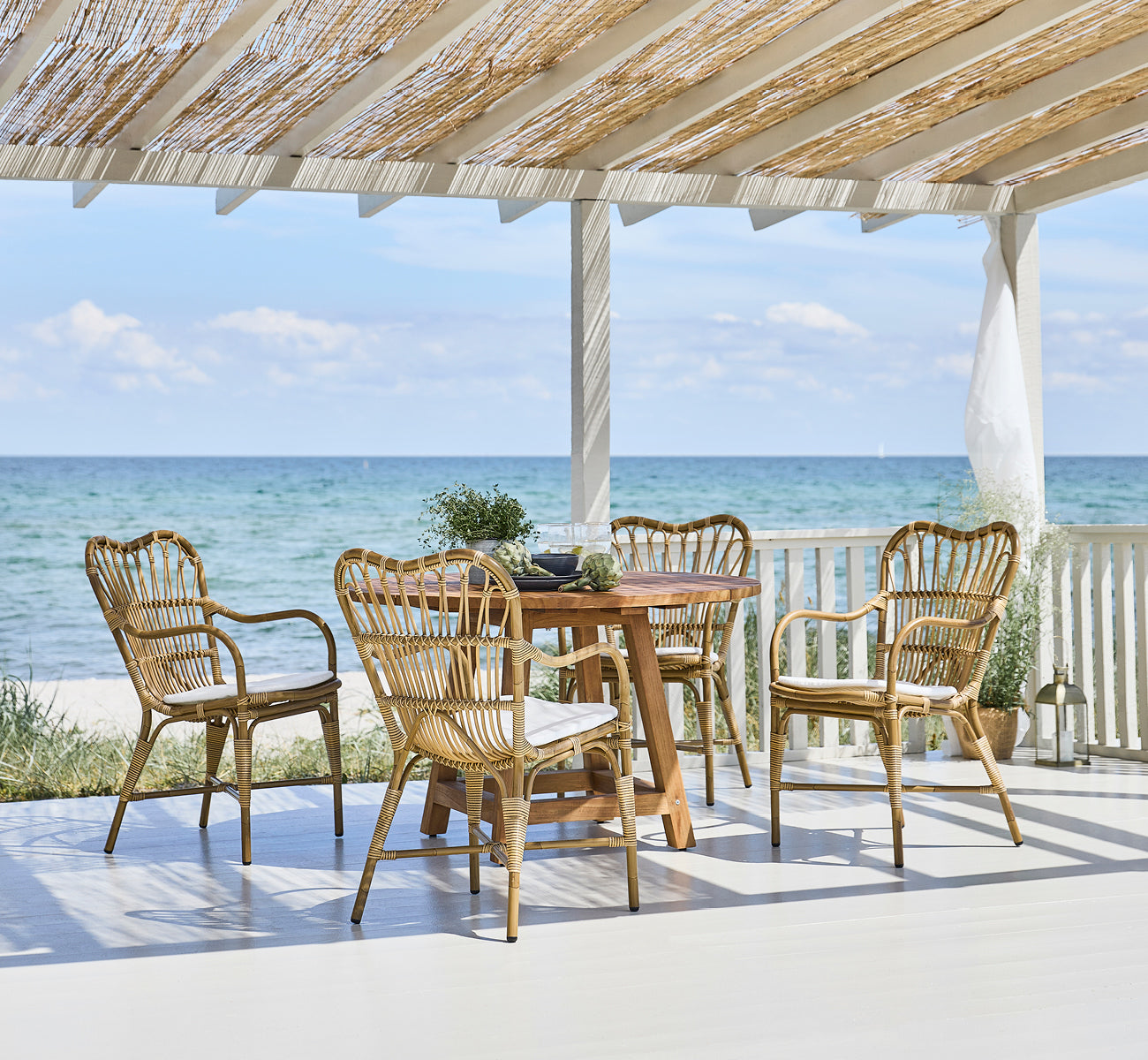 This is a lifestyle image of Margret Exterior Dining Armchair in antique aluminium artfibre. Beach pavilion with teak table and ocean view.