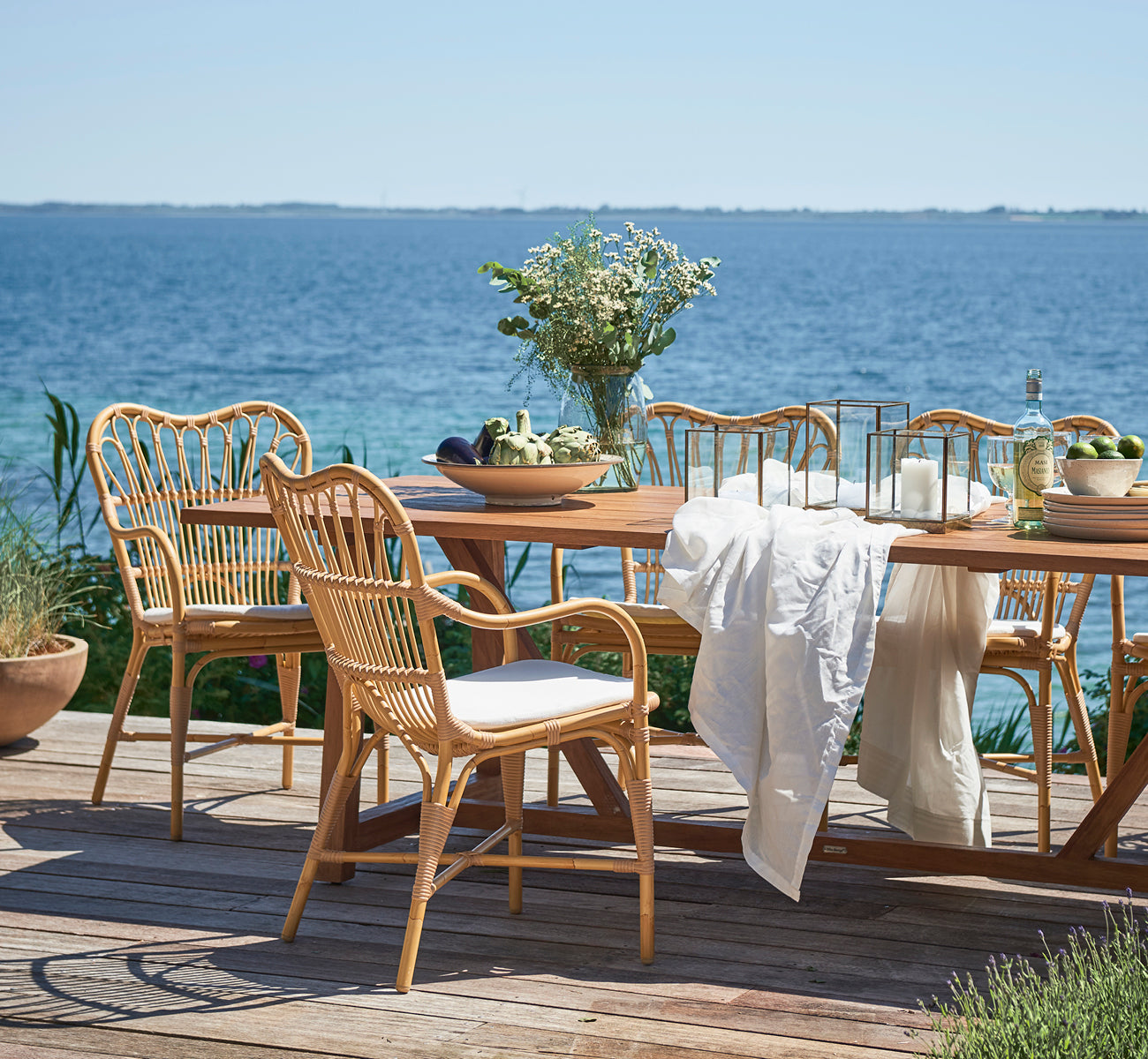 This is a lifestyle image of Margret Exterior Dining Armchair in natural aluminium artfibre. Seaside wooden deck with teak table and flowers.