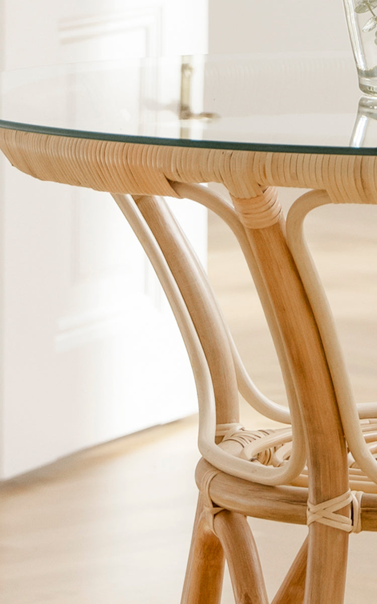This is a close-up image of Tony Café Table in natural rattan. Detail of the round glass top with woven rattan edge and curved legs.