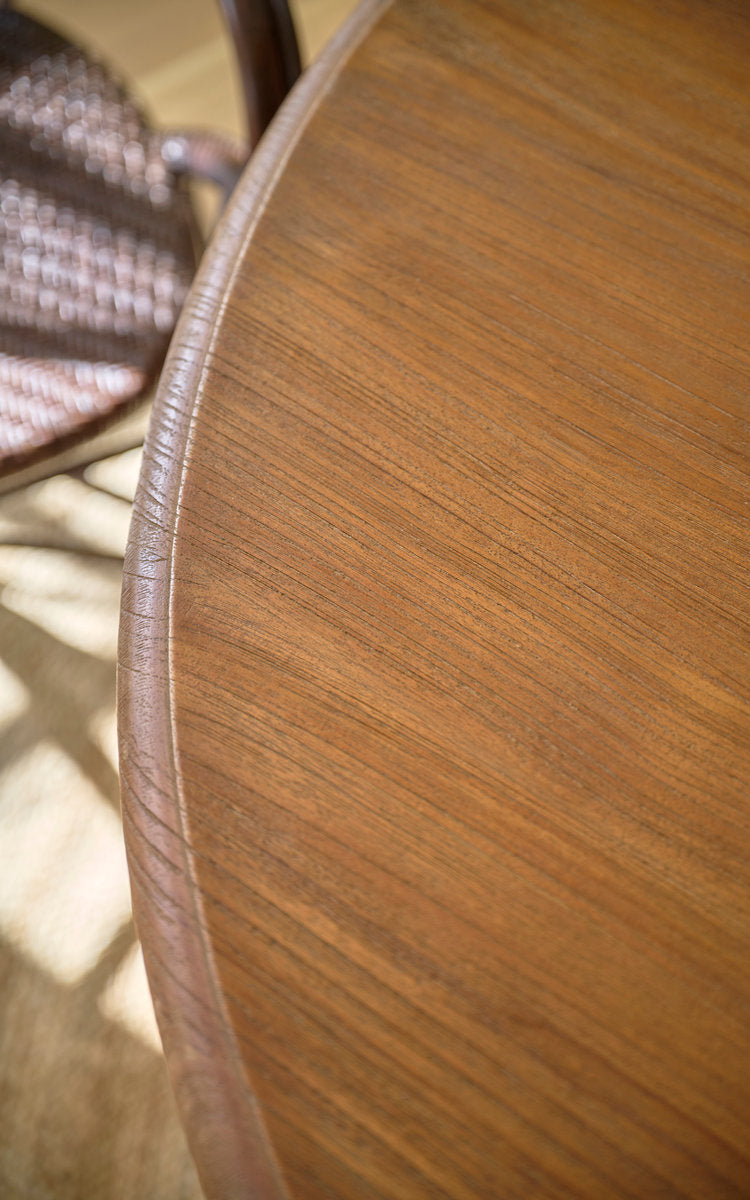 This is a close-up image of Michel Teak Dining Table Ø120 cm in teak indoor. Round teak tabletop edge with rattan chair behind.