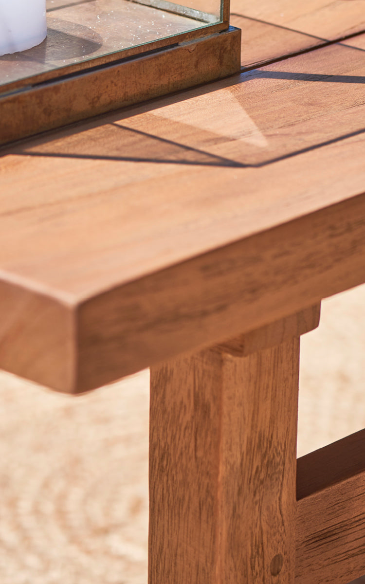 This is a close-up image of Julian Teak Lounge Table 120x120 cm in teak outdoor. The solid teak surface and thick edge are shown in warm sunlight.