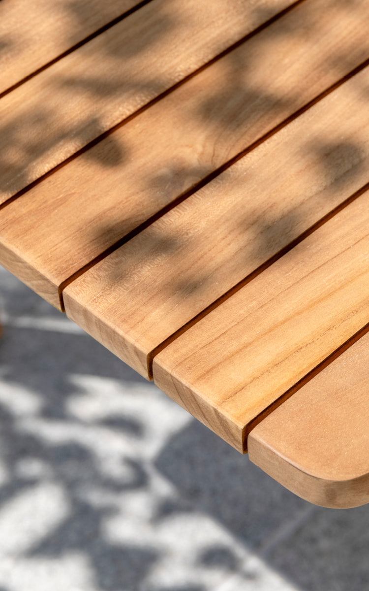 This is a close-up image of Tobias Exterior Teak Table 205x91 cm in teak outdoor. Detail of the teak plank tabletop edge and dappled sunlight.