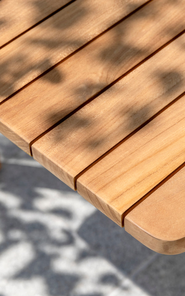 This is a close-up image of Tobias Exterior Teak Table 205x91 cm in teak outdoor. Detail of the teak plank tabletop edge and dappled sunlight.