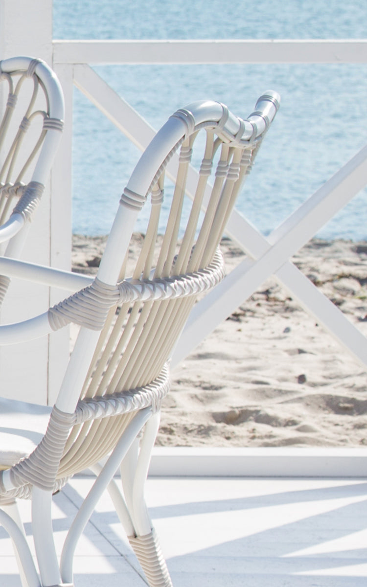 This is a close-up image of Margret Exterior Dining Armchair in dove white aluminium artfibre. White curved spindles with wrapped joints by the sea.