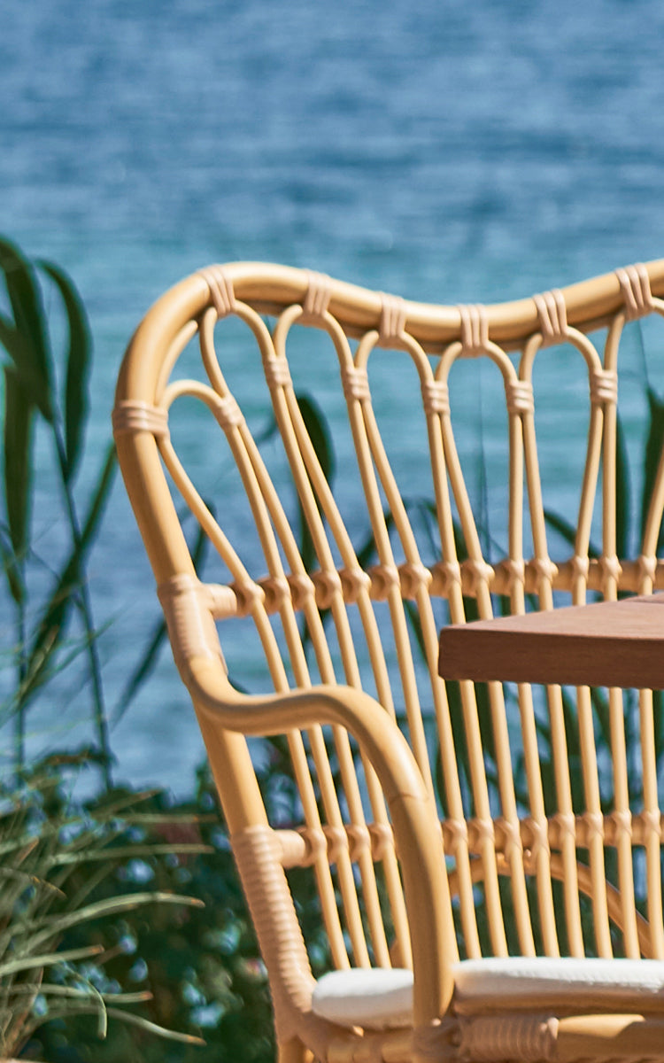 This is a close-up image of Margret Exterior Dining Armchair in natural aluminium artfibre. Curved backrest spindles with ocean view behind.