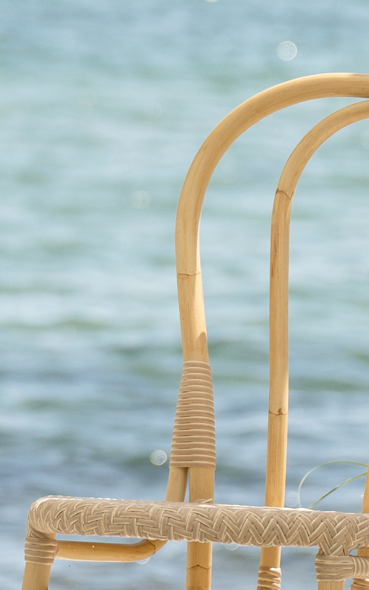 This is a close-up image of Lulu Exterior Dining Chair in natural aluminium artfibre. The curved frame and woven detail are shown by the seaside.