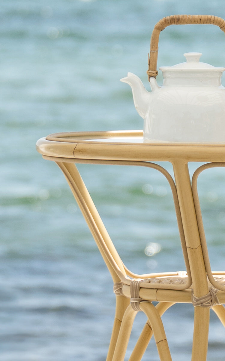 This is a close-up image of Tony Exterior Café Table in natural aluminium artfibre. A white teapot sits on the round top with the sea behind.
