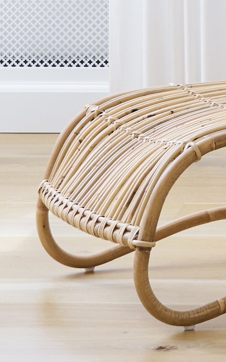 This is a close-up image of Teddy Footstool in polished natural rattan. Detail of the curved rattan rails and plaited wrap of the footstool.