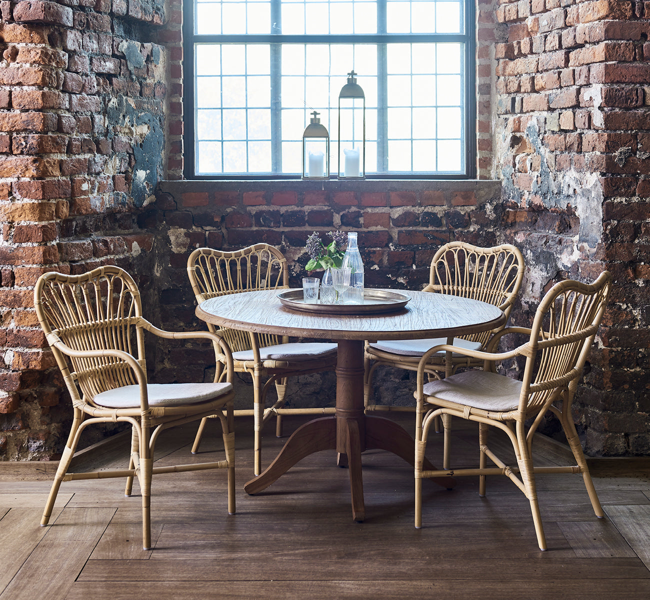 This is a lifestyle image of Margret Dining Chair in natural rattan. Rustic brick interior with round table and lantern on windowsill.