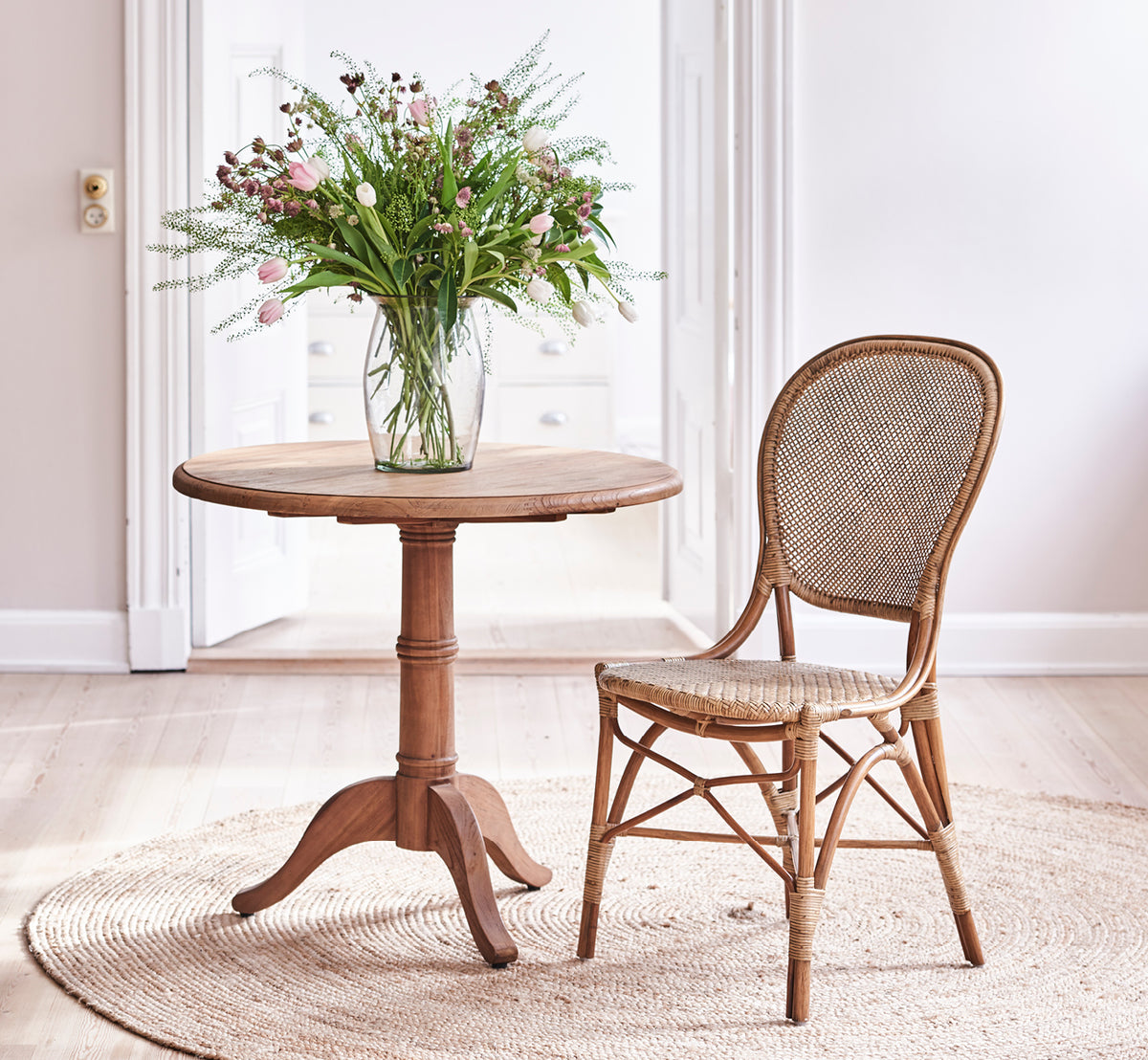 This is a lifestyle image of Rossini Dining Chair in antique rattan. Set around a round teak table on a jute rug in a bright loft.