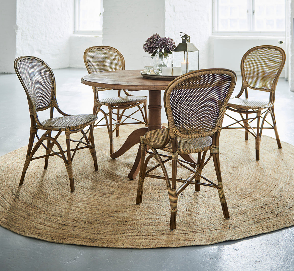 This is a lifestyle image of Rossini Dining Chair in antique rattan. Four chairs around a round teak table on a jute rug in a white loft.