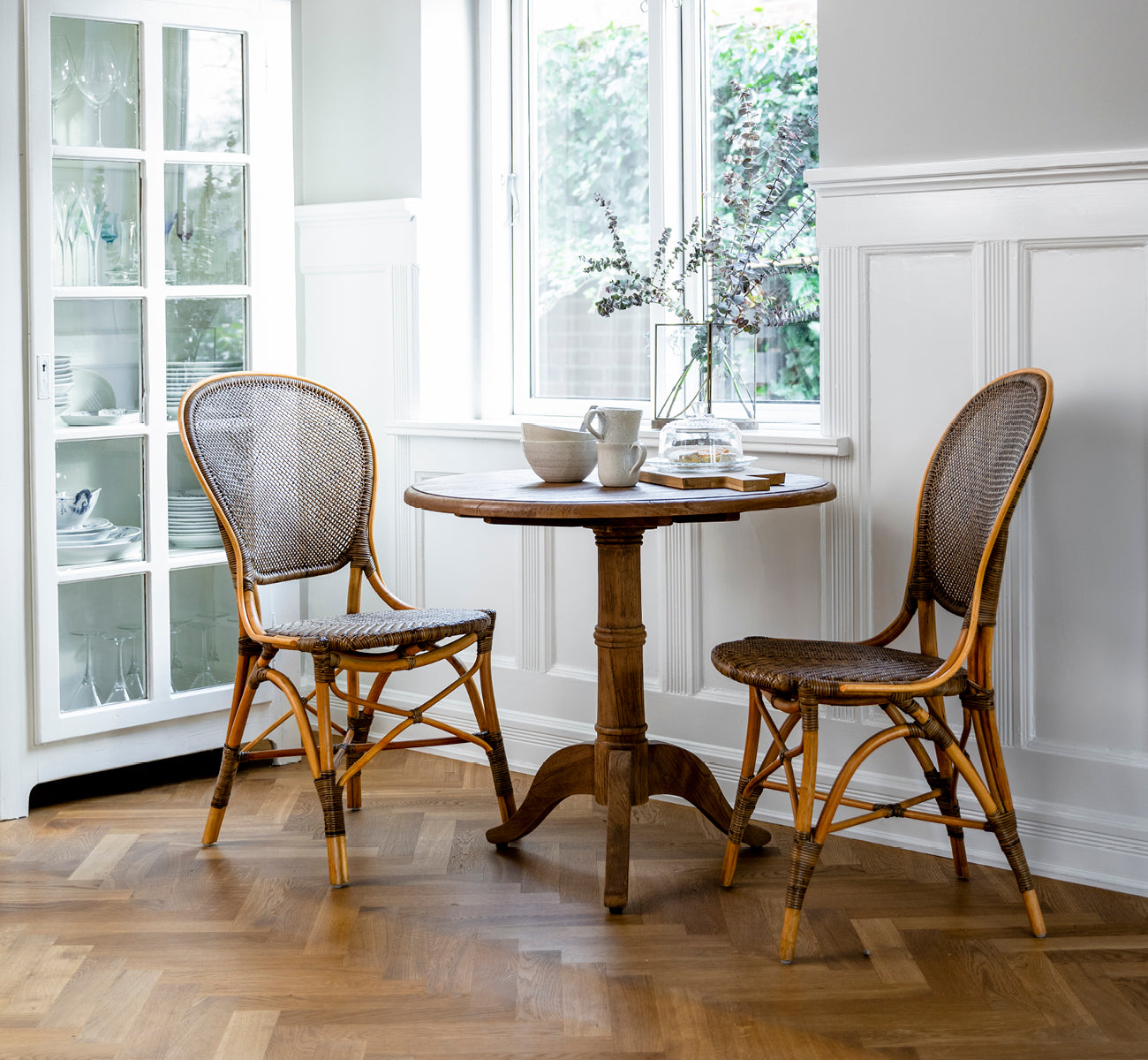 This is a lifestyle image of Rossini Dining Chair in cherry rattan. Two chairs at a small table in a checkered tile courtyard setting.