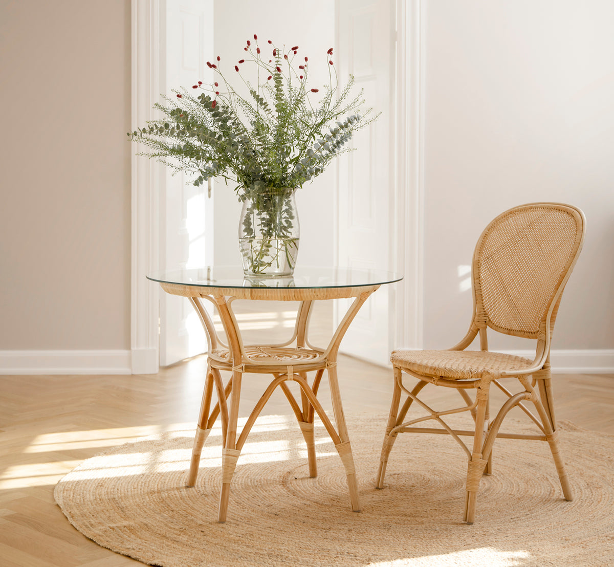 This is a lifestyle image of Rossini Dining Chair in natural rattan. Placed beside a glass-top bistro table on a jute rug in a bright hallway.