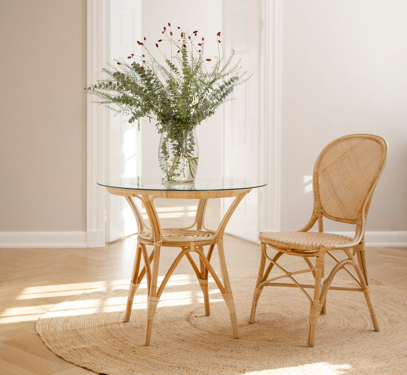 This is a lifestyle image of Rossini Dining Chair in natural rattan. Placed beside a glass-top bistro table on a jute rug in a bright hallway.