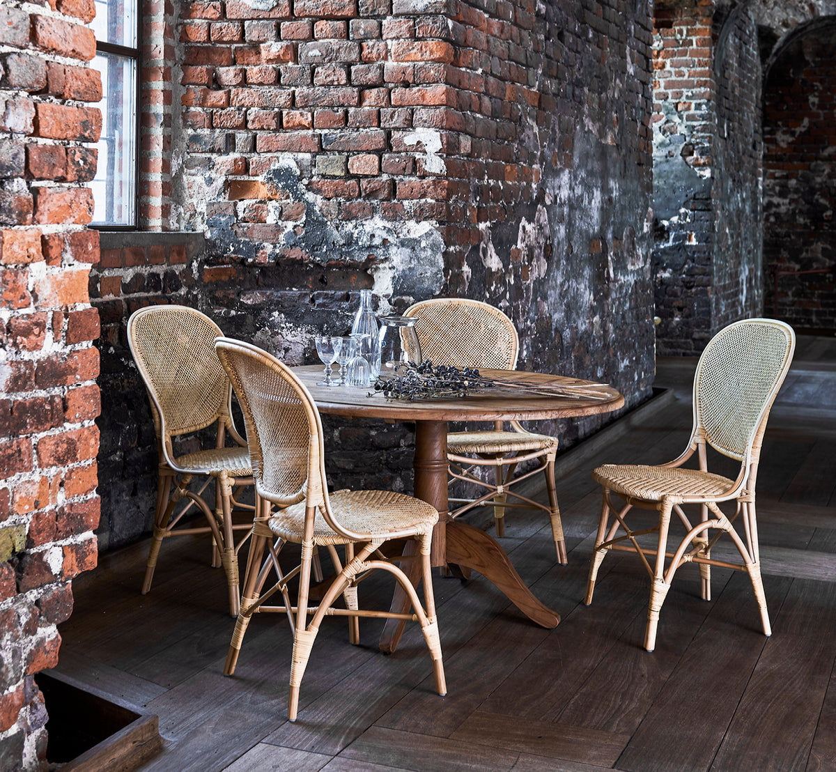 This is a lifestyle image of Rossini Dining Chair in natural rattan. Placed beside a glass-top bistro table on a jute rug in a bright hallway.