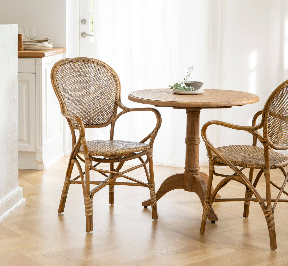 This is a lifestyle image of Rossini Dining Armchair in antique rattan. Paired with a round café table in a light, airy Scandinavian interior.