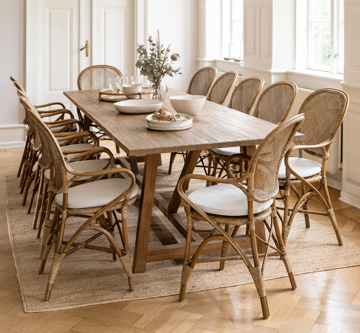 This is a lifestyle image of Rossini Dining Armchair in antique rattan. Surrounding a long teak table with linen draping on a round jute rug.