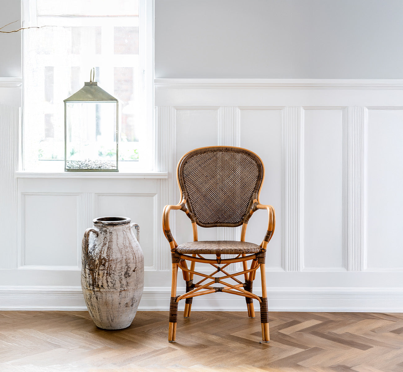 This is a lifestyle image of Rossini Dining Armchair in cherry rattan. Set in a panelled room with herringbone floor beside a rustic urn.