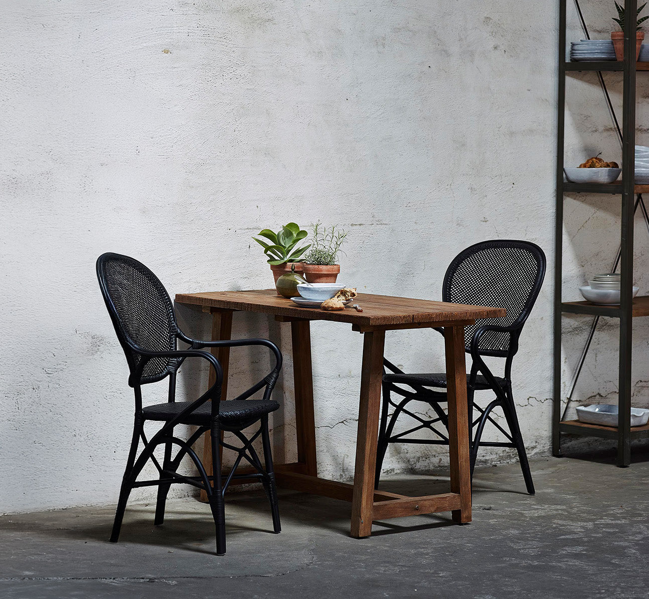 This is a lifestyle image of Rossini Dining Armchair in black rattan. Set around a reclaimed wood table in a raw concrete loft.