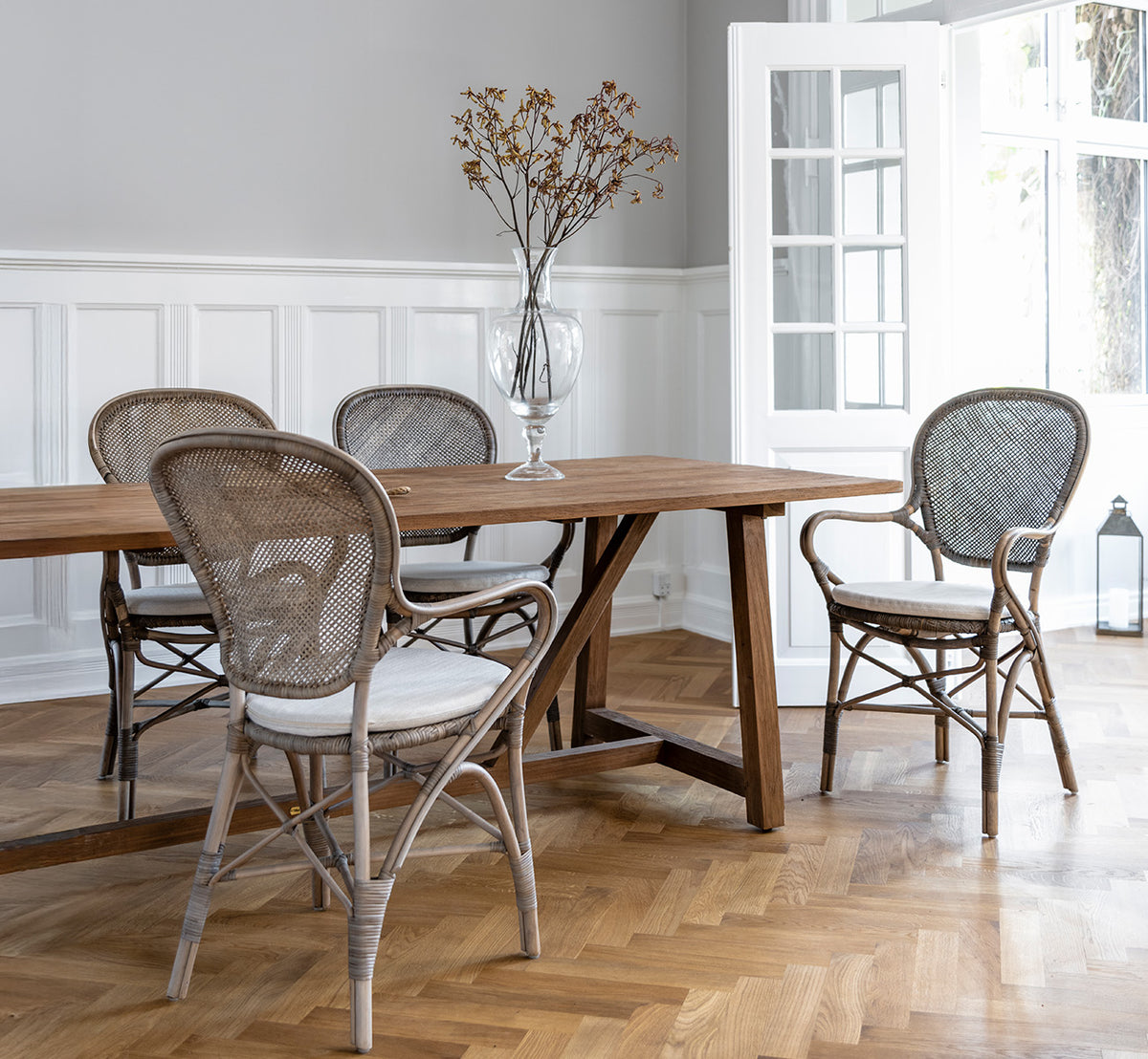 This is a lifestyle image of Rossini Dining Armchair in taupe rattan. Around a wooden dining table on a herringbone parquet floor.