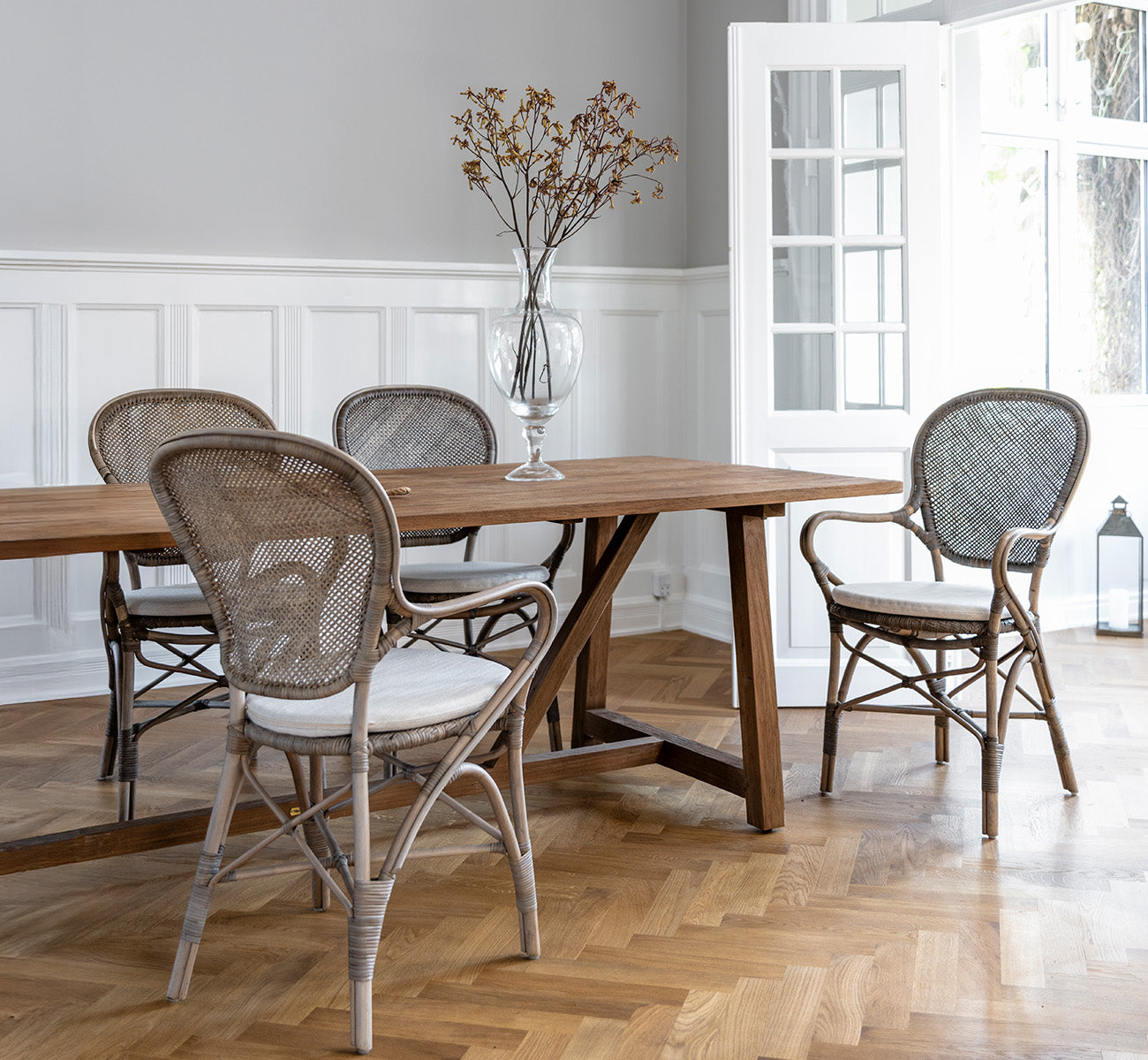This is a lifestyle image of Rossini Dining Armchair in taupe rattan. Around a wooden dining table on a herringbone parquet floor.