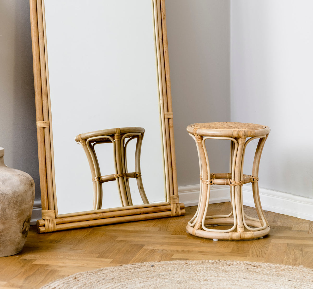 This is a lifestyle image of Bella Stool in natural rattan, in a bright interior with white panelled walls and herringbone parquet.