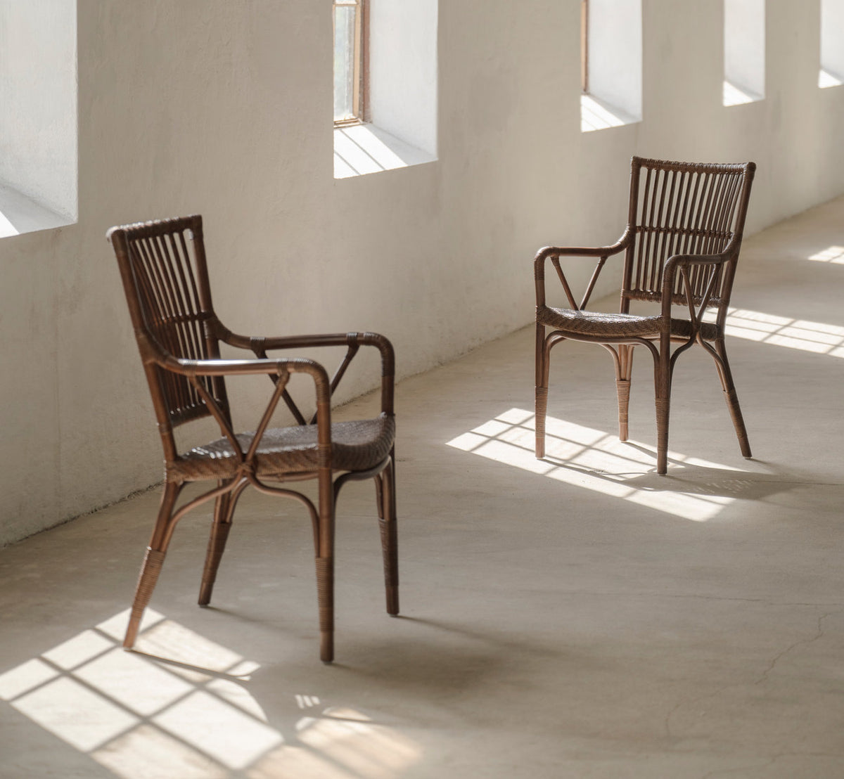 This is a lifestyle image of Piano Dining Chair in brown rattan. Pair in a sunlit room with concrete floor.