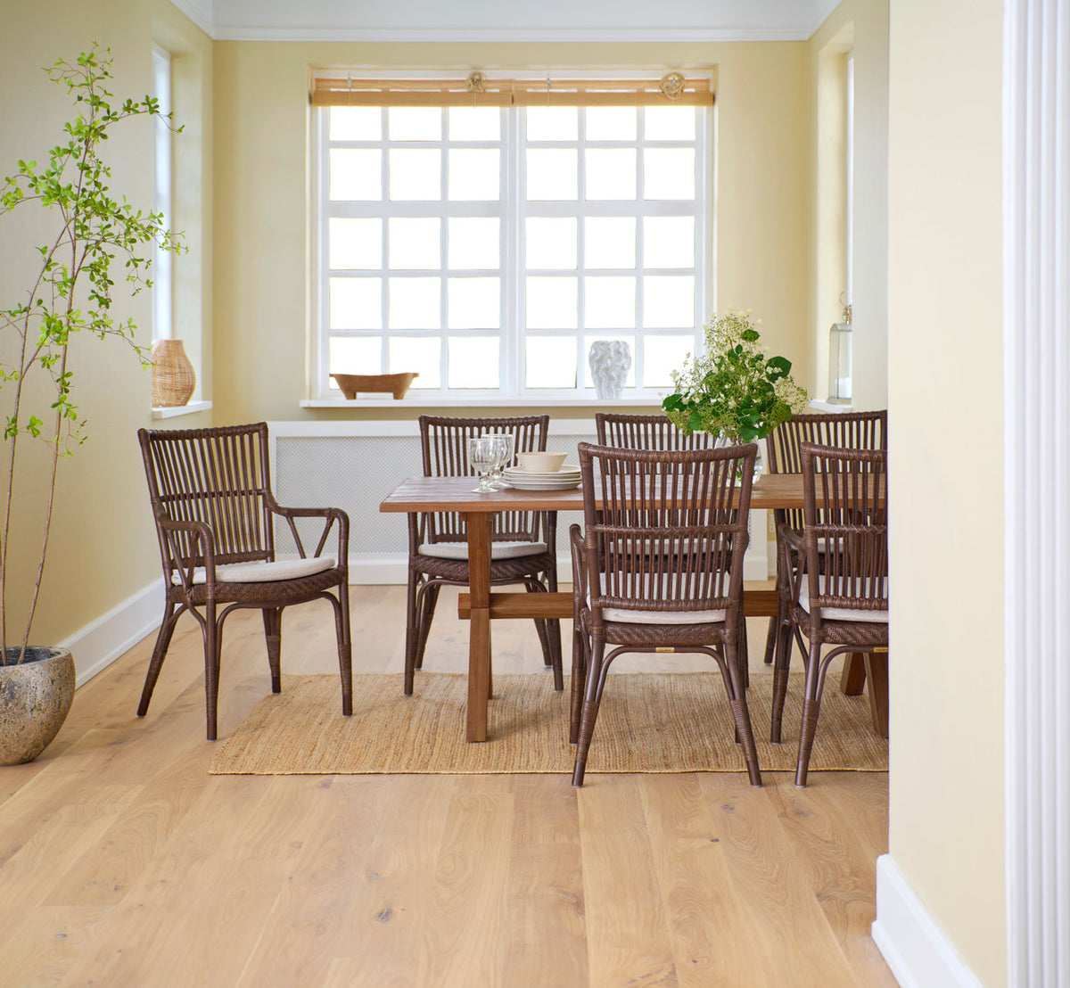 This is a lifestyle image of Piano Dining Chair in brown rattan. Around a teak dining table by a window.