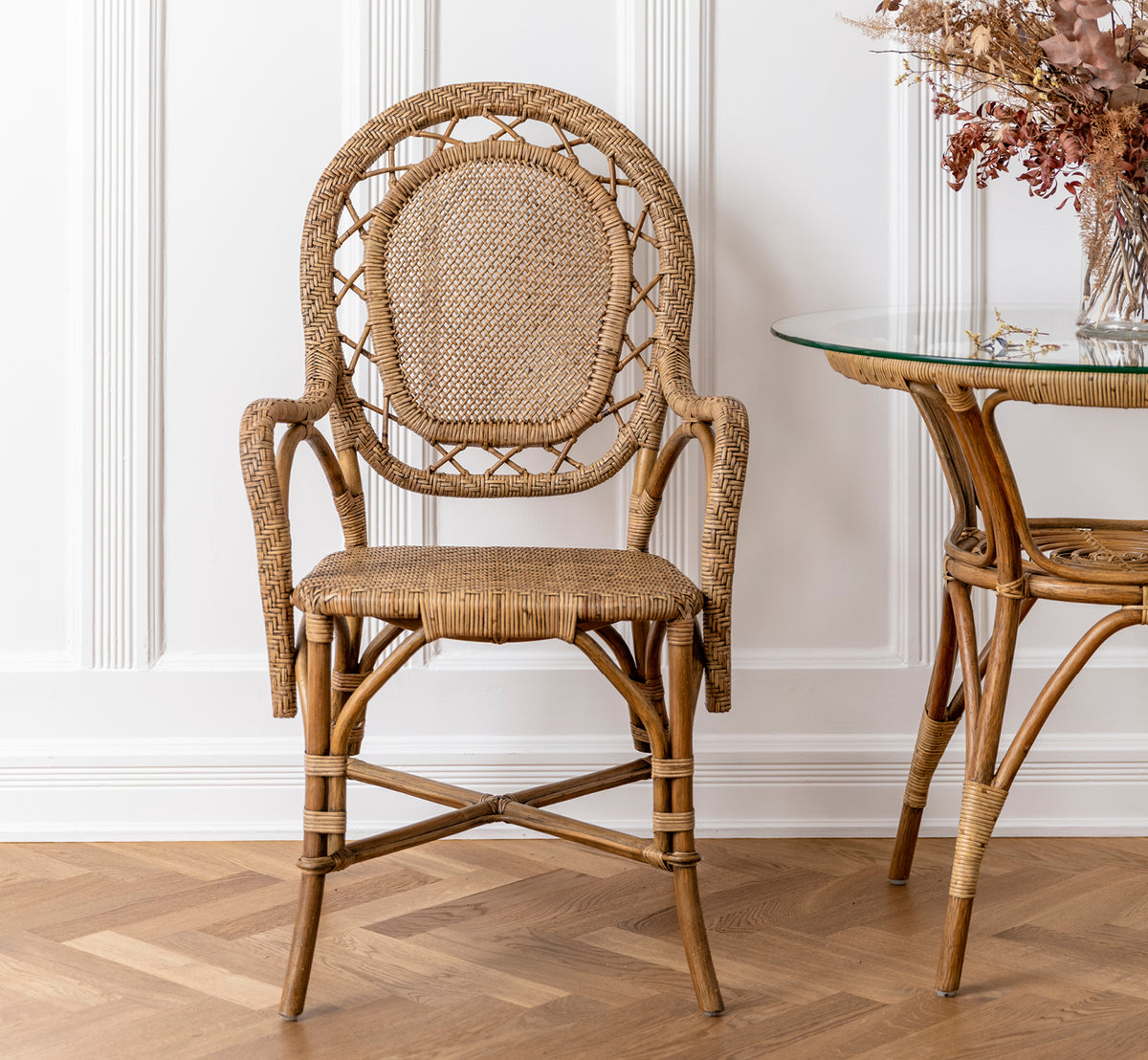 This is a lifestyle image of Romantica Dining Chair in antique rattan. Placed beside a glass rattan table with dried flowers in a paneled room.