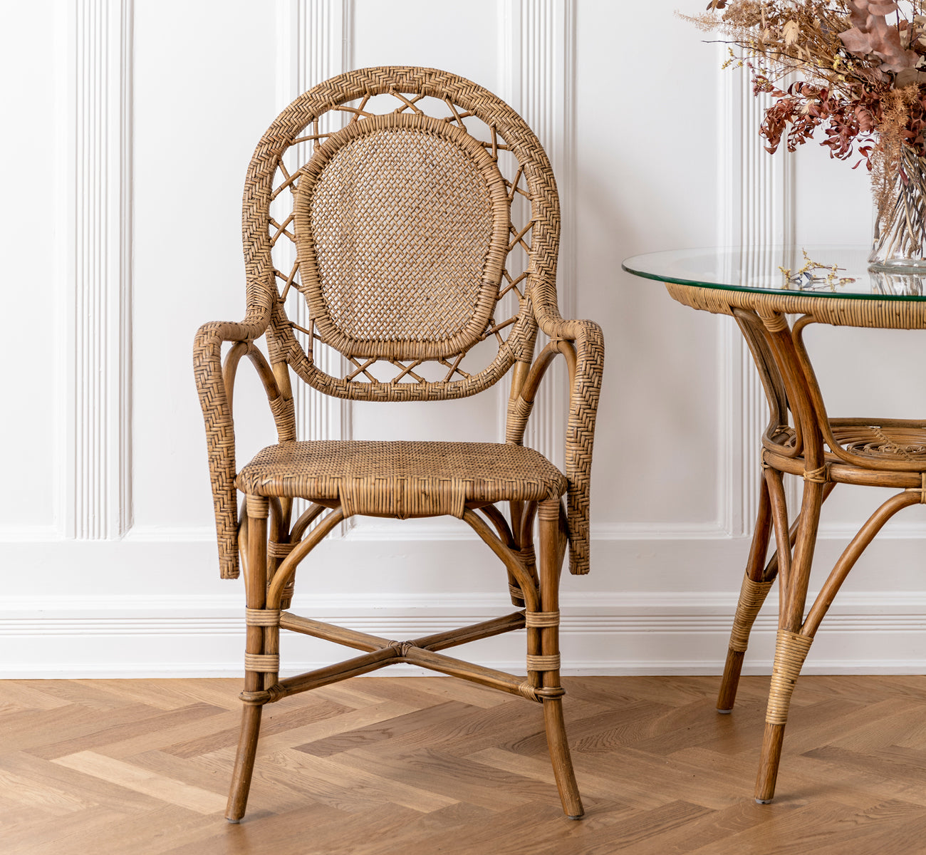 This is a lifestyle image of Romantica Dining Chair in antique rattan. Placed beside a glass rattan table with dried flowers in a paneled room.