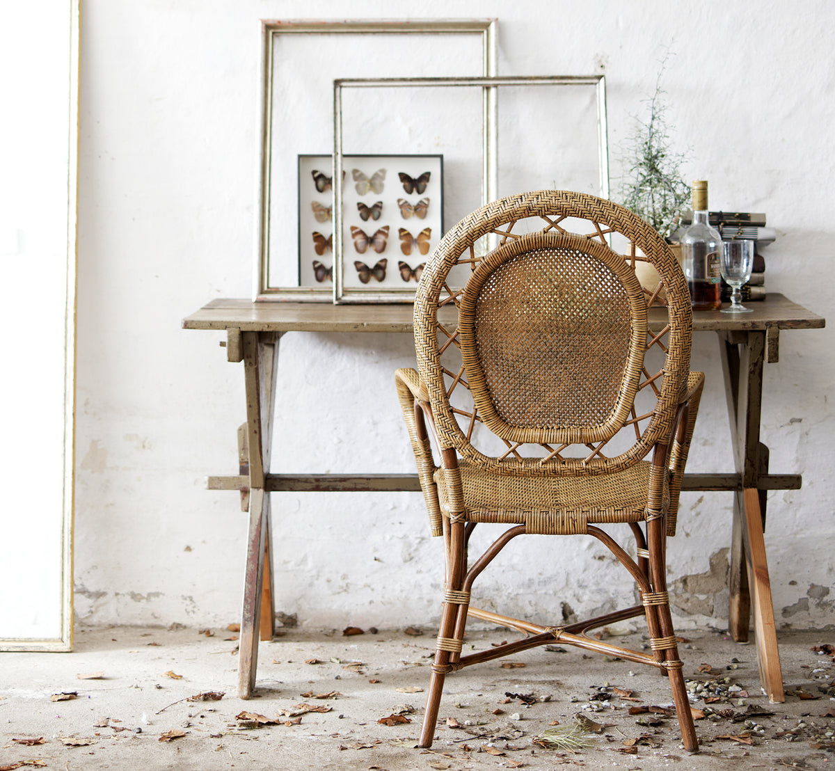 This is a lifestyle image of Romantica Dining Chair in antique rattan. Styled at a rustic wooden table with framed art in a vintage setting.