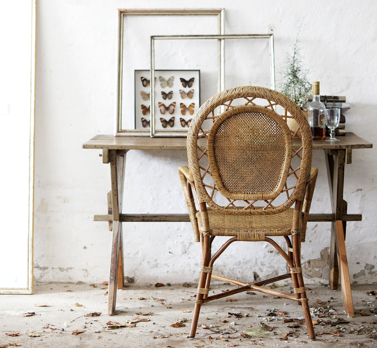 This is a lifestyle image of Romantica Dining Chair in antique rattan. Styled at a rustic wooden table with framed art in a vintage setting.