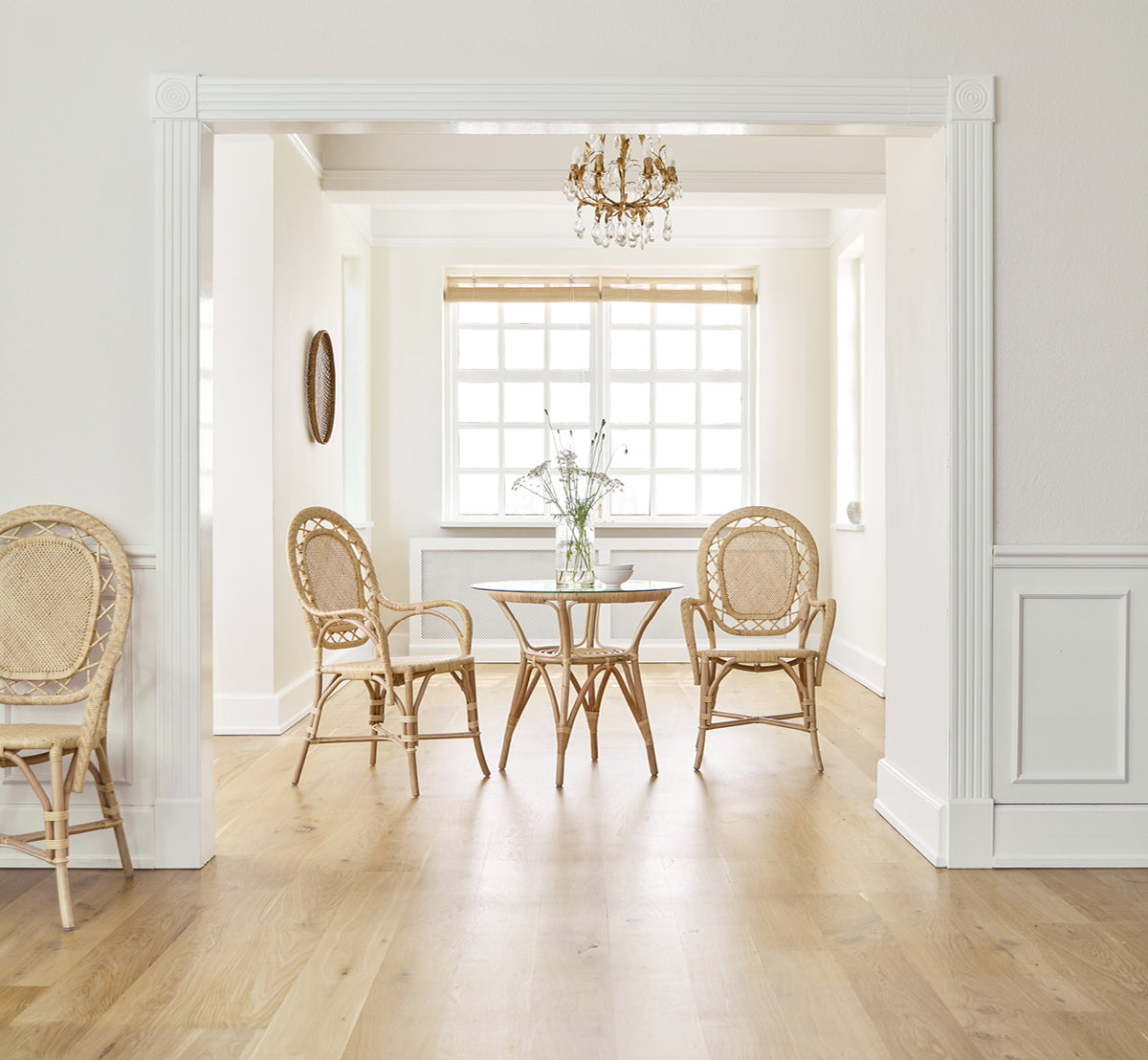 This is a lifestyle image of Romantica Dining Chair in natural rattan. Around a round table in an elegant room with a chandelier and white paneling.