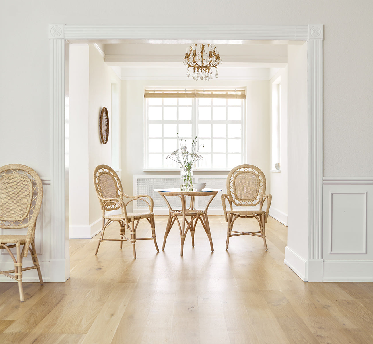 This is a lifestyle image of Romantica Dining Chair in natural rattan. Around a round table in an elegant room with a chandelier and white paneling.