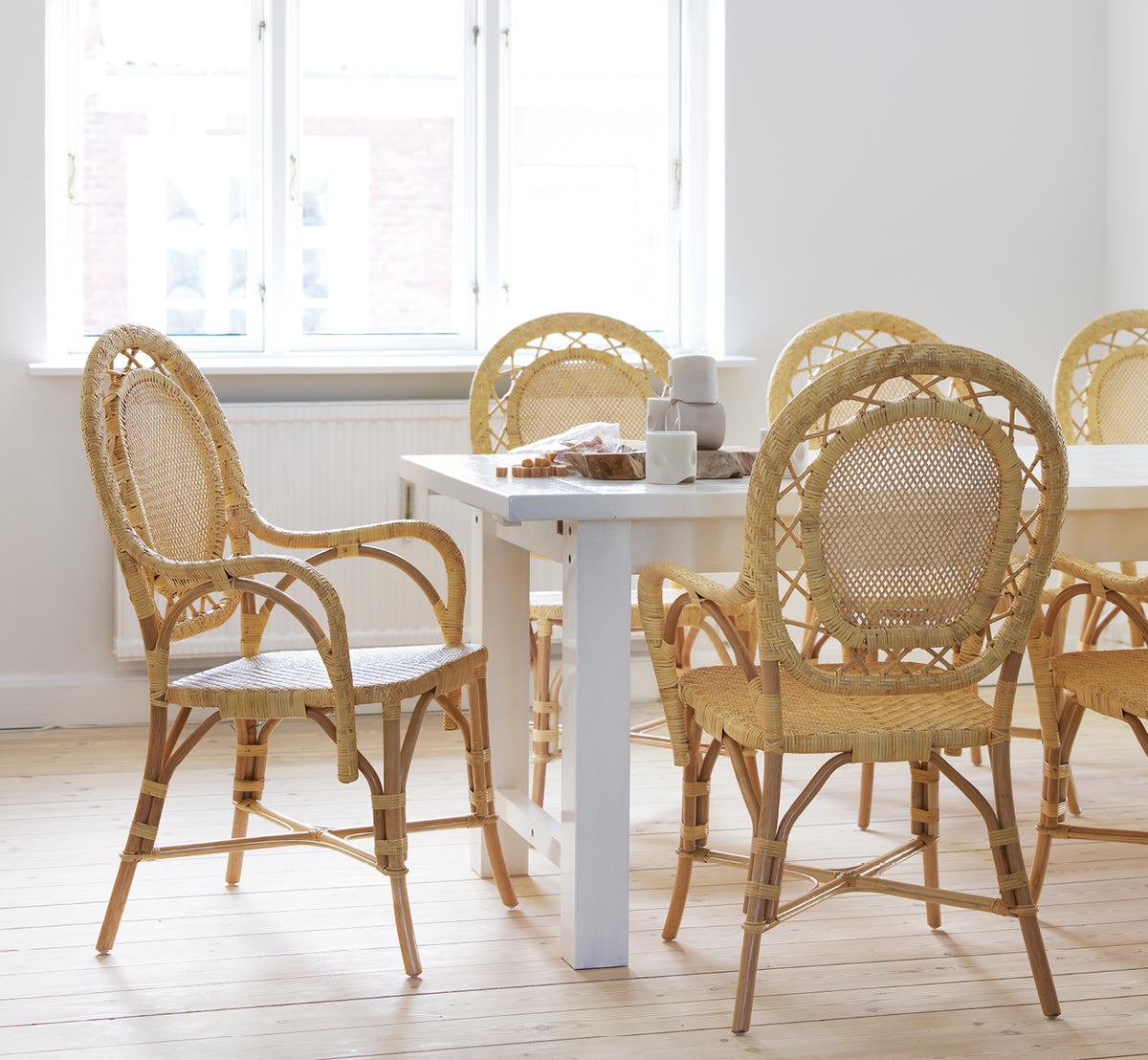 This is a lifestyle image of Romantica Dining Chair in natural rattan. Gathered around a white dining table in a bright Scandinavian room.