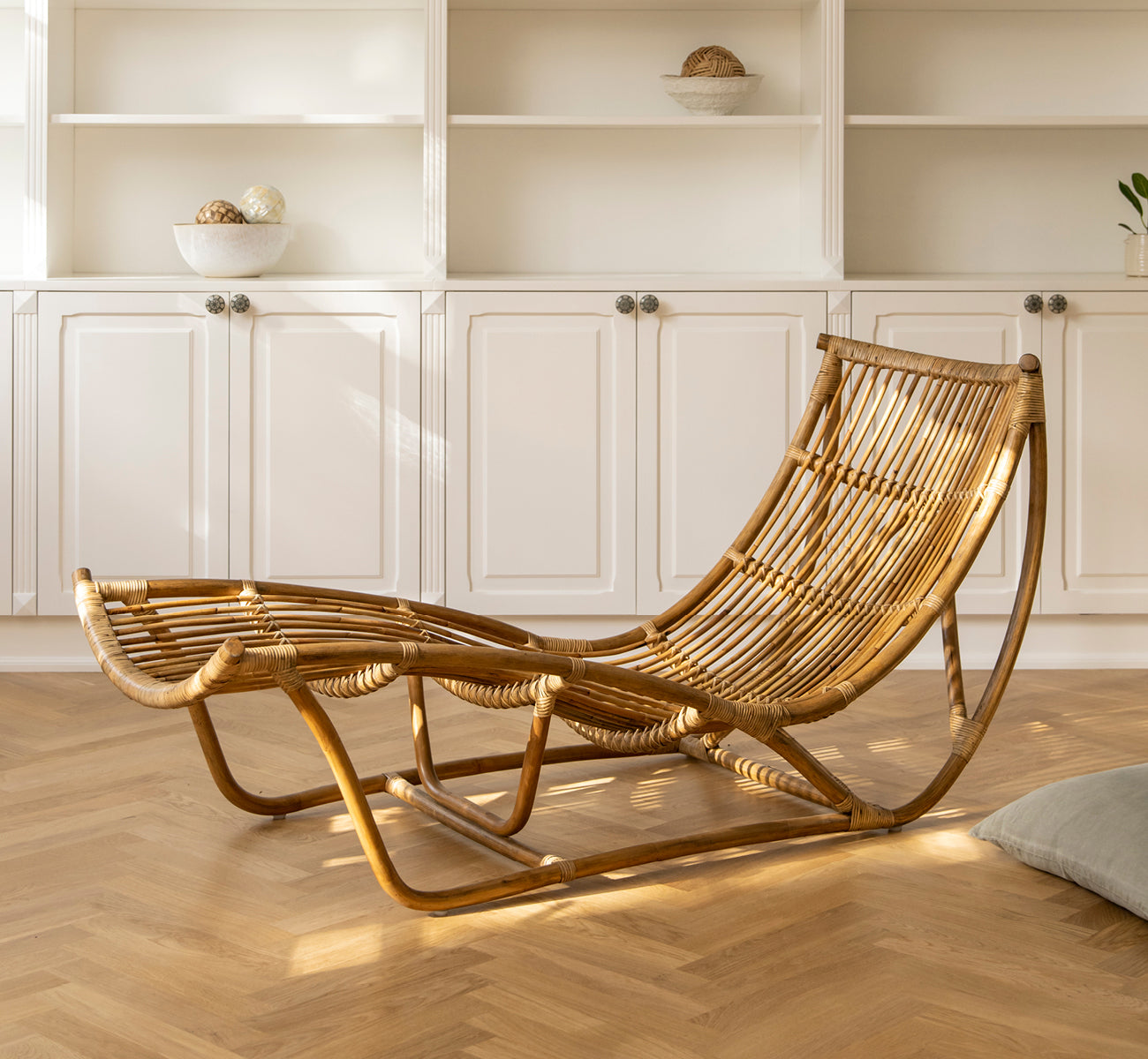 This is a lifestyle image of Michelangelo Daybed in antique rattan. Placed in a sunlit white room with herringbone parquet floor.