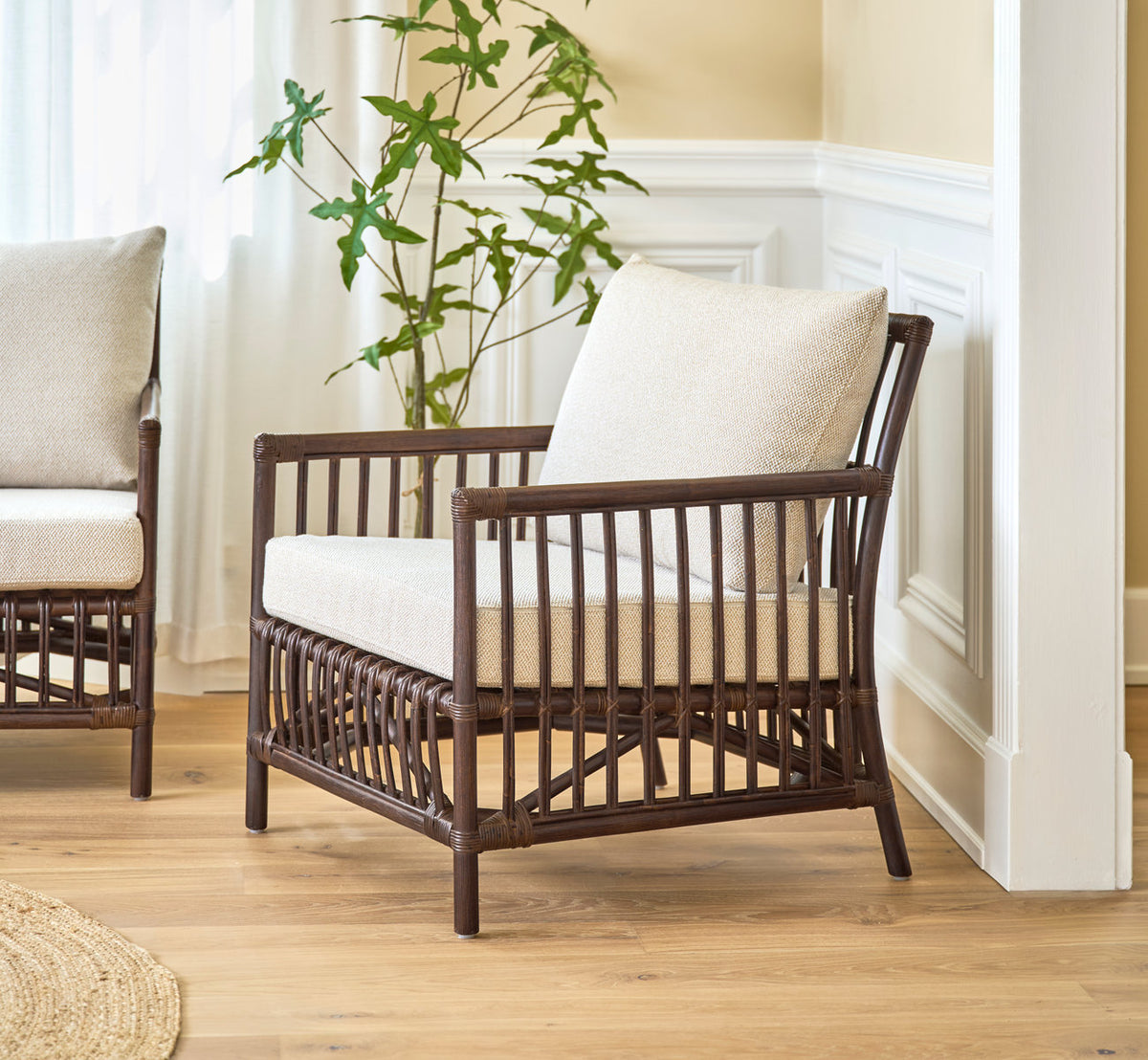 This is a lifestyle image of Caroline Lounge Chair in brown rattan. With linen cushions beside white paneled walls.