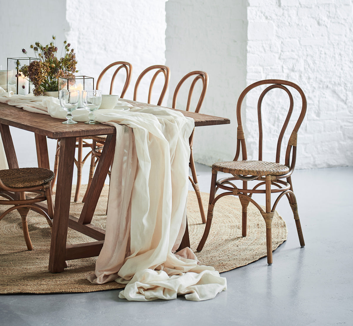 This is a lifestyle image of Lulu Dining Chair in antique rattan. Set at a rustic table with linen draping and candles against a white brick wall.