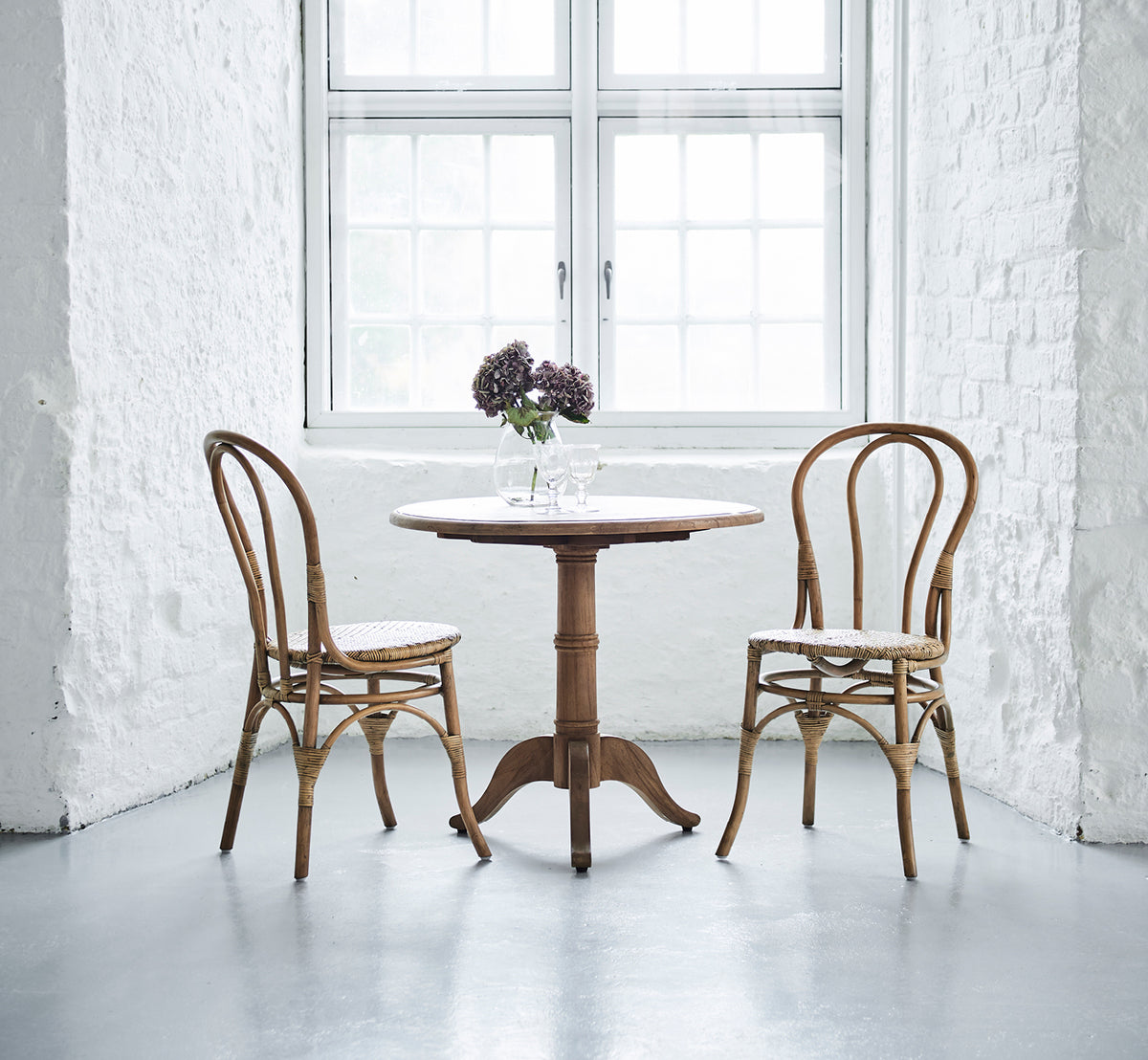 This is a lifestyle image of Lulu Dining Chair in antique rattan. Two chairs flank a round table with flowers in a bright white brick interior.
