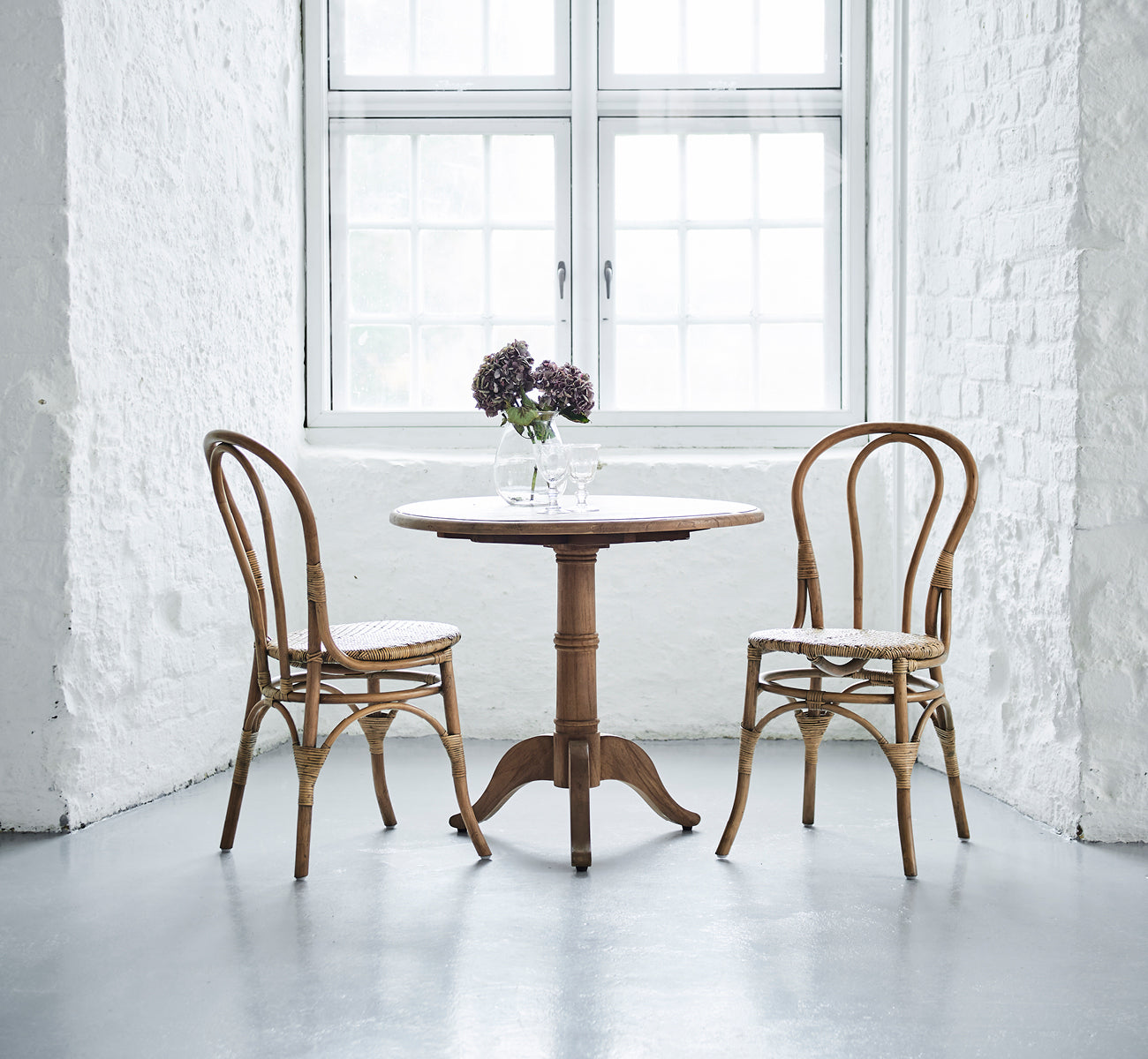 This is a lifestyle image of Lulu Dining Chair in antique rattan. Two chairs flank a round table with flowers in a bright white brick interior.
