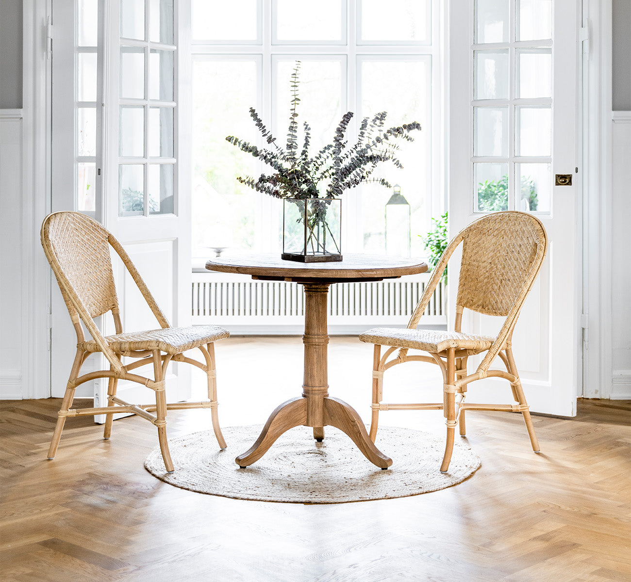 This is a lifestyle image of Alanis Dining Chair in natural rattan, in a bright interior with white panelled walls and herringbone parquet.