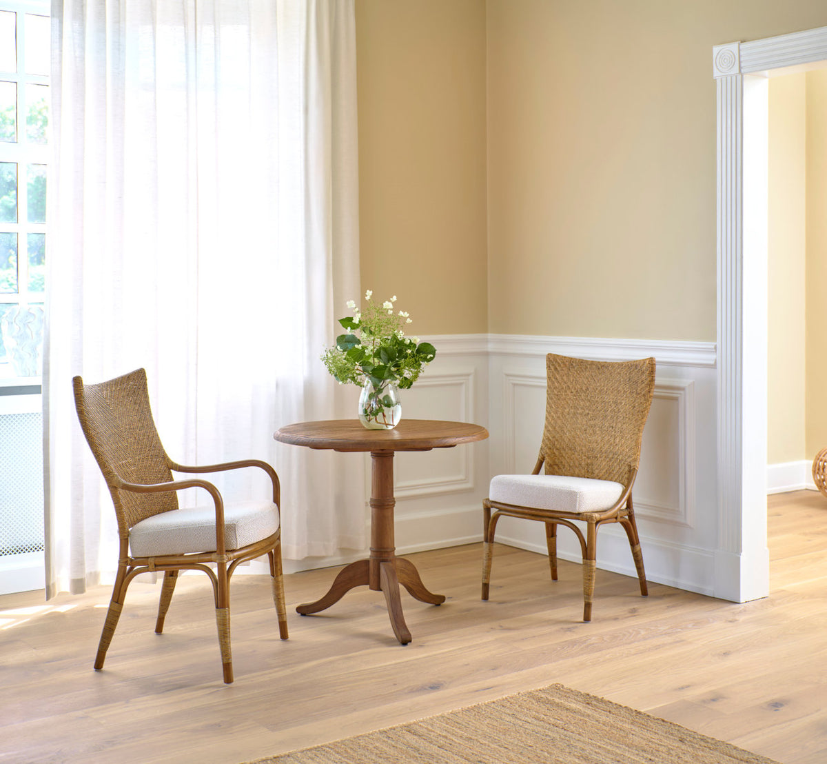 This is a lifestyle image of Melody Dining Chair in antique rattan. Pair at a teak café table by white panels.