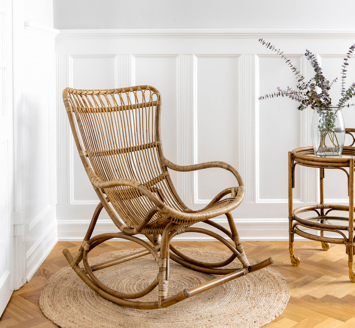 This is a lifestyle image of Monet Rocking Chair in antique rattan. In a butterfly wallpaper room with glass-top rattan side table.