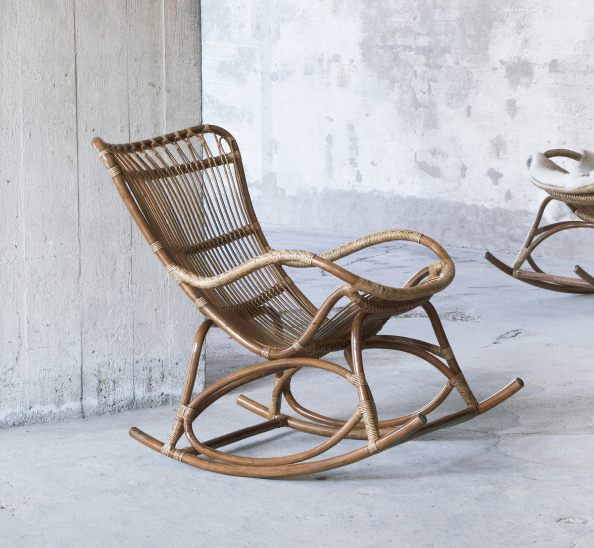 This is a lifestyle image of Monet Rocking Chair in antique rattan. In a rustic concrete room with another rocker in the background.