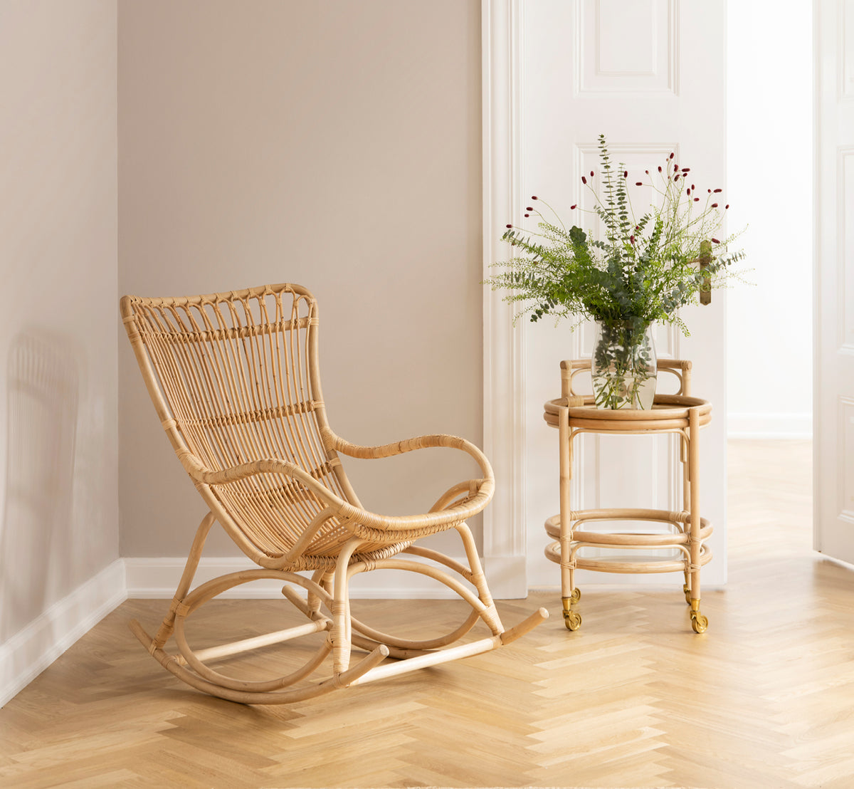 This is a lifestyle image of Monet Rocking Chair in natural rattan. In a cozy corner with herringbone floor and rattan trolley.