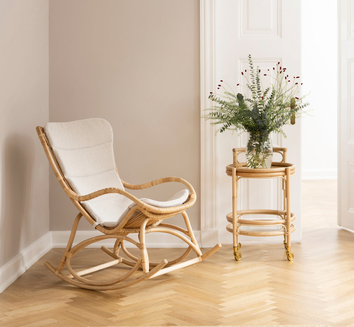 This is a lifestyle image of Monet Rocking Chair in natural rattan. In a blush-walled room with herringbone parquet and eucalyptus.